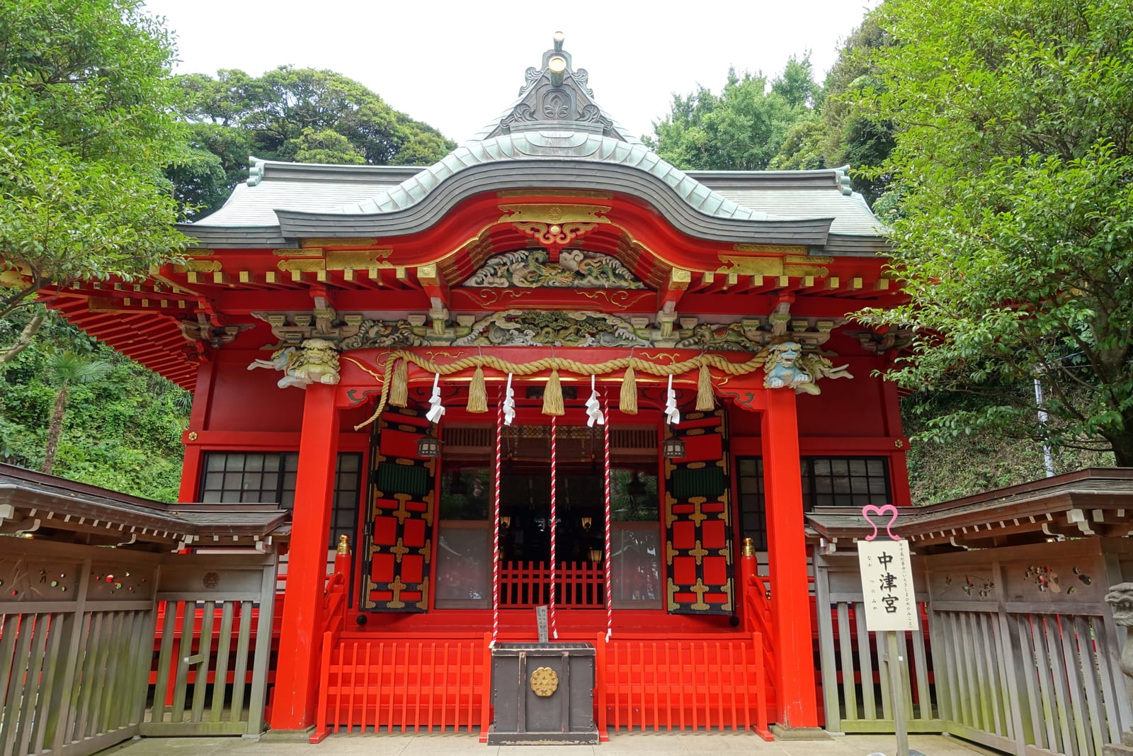 Um edifício de santuário vermelho e caixa de oferendas em Nakatsu-no-miya, emoldurado por árvores verdes.